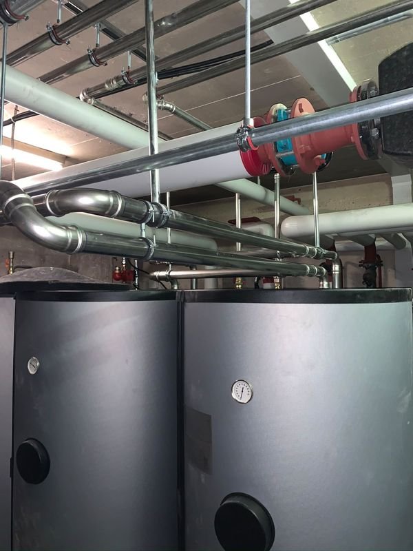 Deux ballons de stockage gris avec thermomètre, raccordés par des conduites en inox serties suspendues au plafond du local technique.