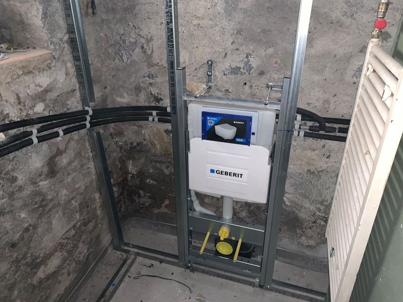 Bâti-support Geberit pour WC suspendu, fixé sur une ossature métallique contre un mur en pierre, avec les conduites d'eau sous gaine raccordées.