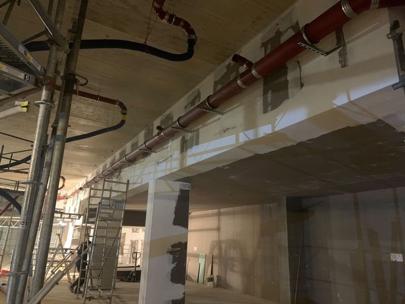 Chantier neuf : conduite rouge suspendue sous la dalle sur des consoles, avec des piquages en attente, échafaudages autour.