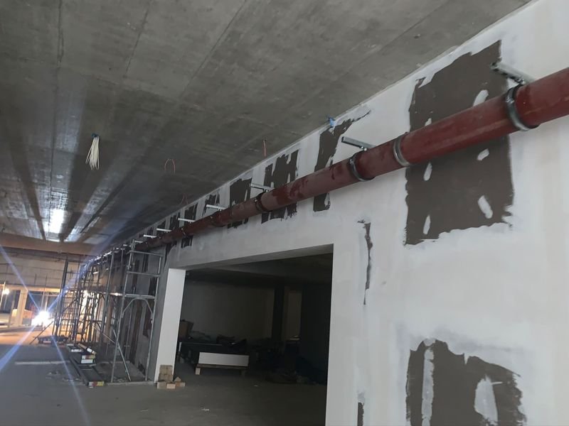 Chantier neuf en sous-sol : longue conduite rouge suspendue en haut d'un mur, fixée par des colliers sur des consoles métalliques.