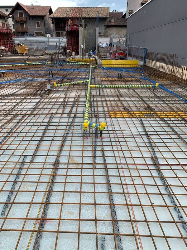 Chantier neuf : conduites protégées par un ruban jaune et bleu, bouchonnées à leurs extrémités et fixées dans le ferraillage d'une dalle, avant le coulage du béton.